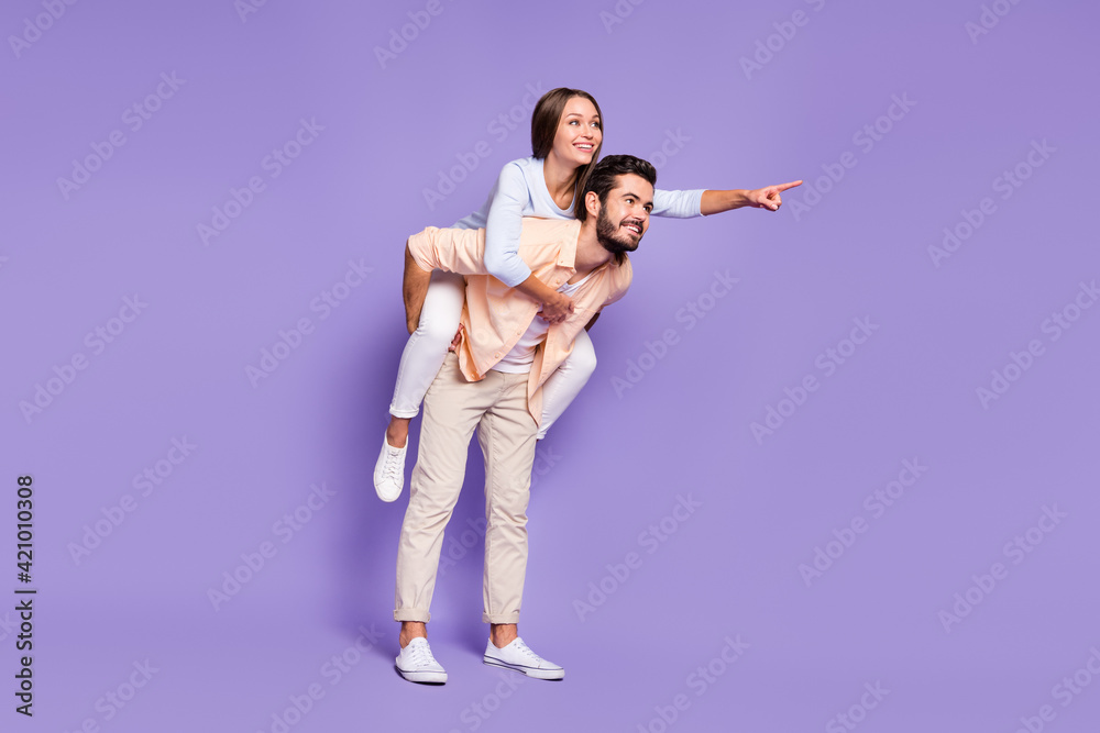 Poster Full size photo of happy young couple man piggyback woman point finger empty space isolated on purple color background