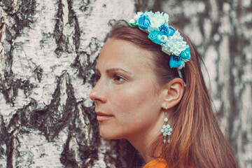 Smiling young woman wearing a decorative floral headband is looking at camera. Close-up portrait of a natural girl without makeup over a birch bark background. Spring is coming.