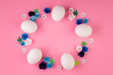 White Easter eggs and paper flowers in blue tones are placed on a pink background.