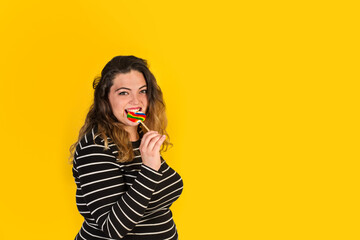 Mujer joven y sonriente mordiendo un lollipop de colores sobre un fondo amarillo. Vista de frente y de cerca. Copy space