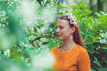 Smiling pretty young woman wearing a decorative floral headband is looking down away. Close-up portrait of a natural girl without makeup is enjoying the smell in blooming garden. Spring is coming.