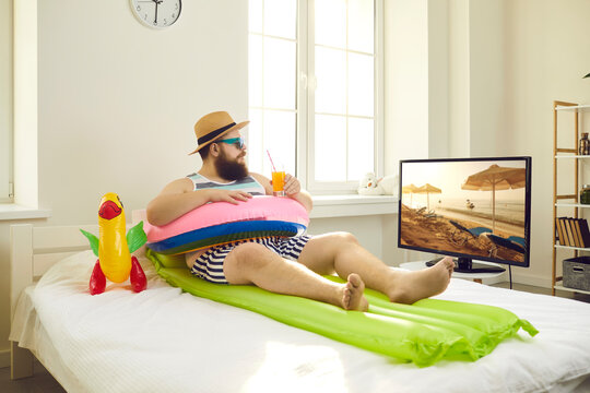 Funny young man sitting on bed with inflatable swim ring, sipping cocktail and watching travel show on TV. Concept of canceled summer holiday plans, vacation in lockdown at home or Covid-19 quarantine