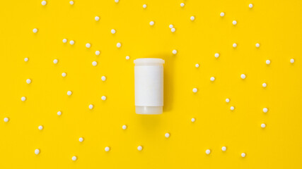 A white jar with an empty label and a place for the inscription lies in the center of the yellow background, surrounded by randomly placed small homeopathic granules.