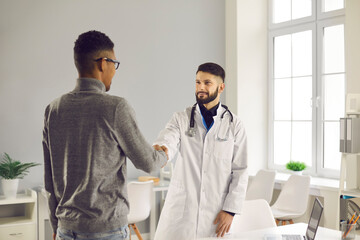 Smiling man doctor therapist shaking hand to black man patient after successful checkup consultation in medical clinic office. Healthcare, medicare, communication with doctor concept