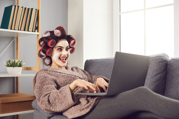 Funny crazy cheerful woman distant worker in curlers and cosmetics mask on face sitting on sofa and working online at laptop from home. Funny women working at home concept