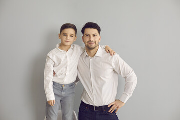 Positive father and son in stylish white shirts and jeans standing hugging and looking at camera over grey wall background. Happy fatherhood, childhood, family concept