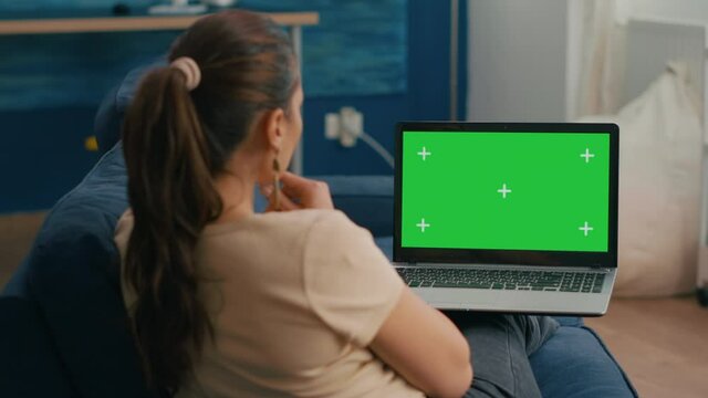 Freelancer woman sitting on sofa in home office typing on laptop computer with mock up green screen chroma key display. Caucasian woman working on financial graphs using isolated pc