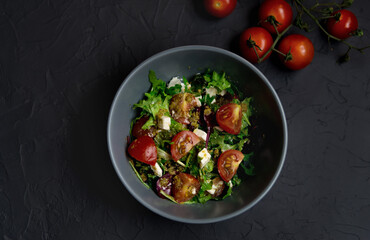 Fresh salad with tomatoes, arugula and cheese cubes on black plate on Dark grey black slate background