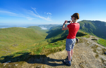 Naklejka premium A female hiker taking photos down the glen of Allt a Chobhair from the mountain summit of Beinn Ghlas in the Scottish Highlands, UK landscape.