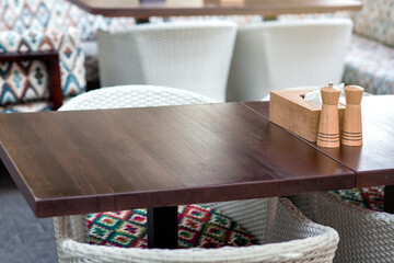 wooden napkin holder with salt and pepper shaker on a wooden table of a summer terrace of a restaurant with wicker chairs, close-up empty brown tabletop surface nobody.