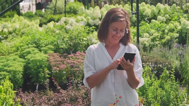 Medium shot of cheerful caucasian woman admiring beautiful green plants and flowers in summer yard under sunlight, taking photos of them on her smartphone
