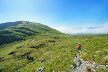 A female hiker and their dog walking down from the mountain summit of Meall Corranaich with Beinn Ghlas in the distance in the Scottish Highlands, UK landscape.