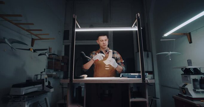 Wide angle male caucasian woodworker, craftsman painting wing detail of wooden bird toy with white paint roller in workshop at night. Man wearing checkered shirt and orange woodworking aprons