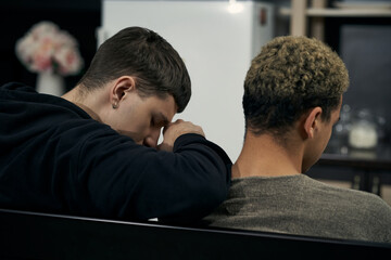 Staged photo illustrates problems and conflicts in gay couple relationships. Moment of showdown: two young men are sitting in the kitchen. Black-haired man's leant on partner's shoulder.