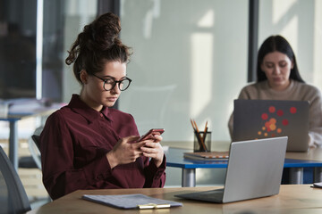 Young businesswoman in eyelasses sitting at her workplace with laptop and using mobile phone