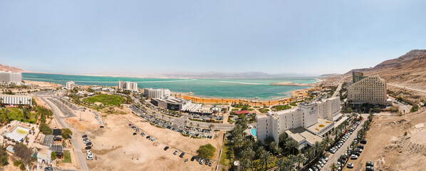 Plakat Aerial panoramic view from a drone of beach on Ein Bokek embankment on coast of Dead Sea, tourist hotels and car parks, the sea itself and the mountains of Jordan visible in the distance, in Israel