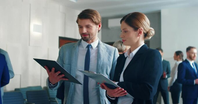 Two business colleagues, man and woman, stand in the middle of hall and discuss conference, sharing their impressions and notes.