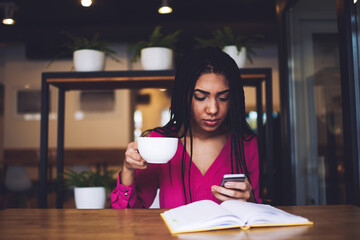 Wistful black woman drinking coffee and using smartphone