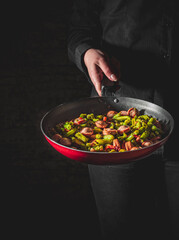 Chef cooking sausages with green beans in pan on black background
