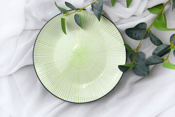 Empty ceramic plate with eucalyptus branch on white silk background. Mock up elegant tableware composition. Summertime minimalist concept for wedding invitation, restaurant menu or spa branding.