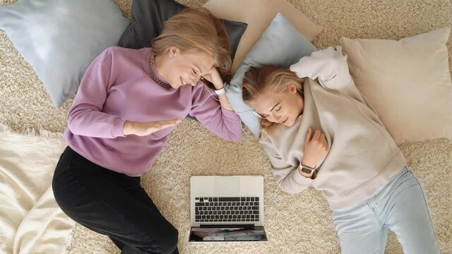 Two young cute sisters lying on the floor on pillows at home and watching funny movie on laptop