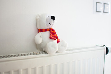 White teddy bear sitting on central heating radiator. Energy saving and save money concept with copy space.