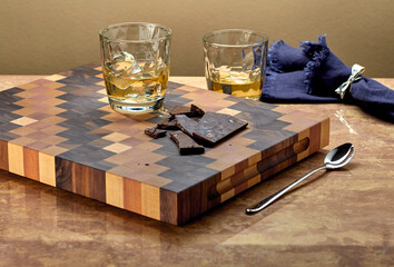 Two glasses of whiskey on the rocks sit on a beautiful wooden chopping board, with chocolate slices ready to eat. Horizontal position.