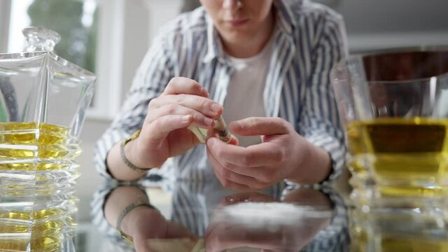 Unrecognizable young man rolling dollar banknote with alcohol and white powder on glass table at front. Addicted Caucasian millennial drinking and sniffing drugs indoors. Addiction concept.