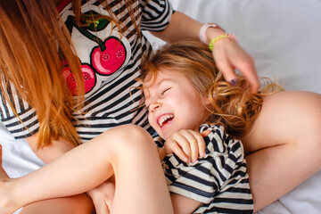 Mom and daughter in matching striped clothes sit on the bed in the white bedroom. Daughter laughs