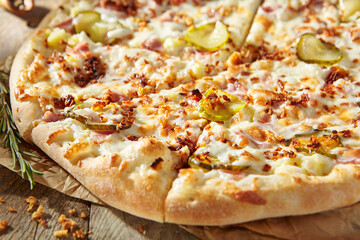 Tasty pizza with ham, gherkins, fried onions and pickle cucumber. Garnished on parchment with pizza ingredients on wooden table. Rustic style, sunlight.