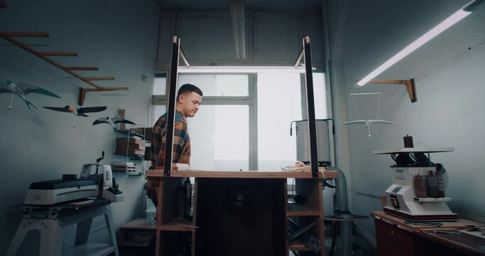 Wide shot handsome male craftsman turns on light enters small woodworking workshop and starts process of creating beautiful wooden bird eco toys. Manufacturing, small business and hobby concept