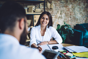 Portrait of cheerful female journalist in smart casual clothing smiling at camera during collaborative meeting with male colleague, happy Caucasian woman posing while listening ideas on meeting