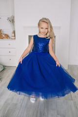 Little beautiful girl in a blue dress stands in a room near the fireplace light interior