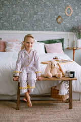 Child girl sitting in pajamas in the bedroom Blonde with blue eyes next to a toy beige plush hare a cup with cocoa and a candle Stylish interior Model portrait