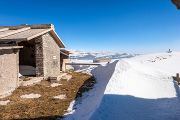 Typical old stone cow shed on Lessinia Plateau (Altopiano della Lessinia) in winter with snow near the peak of Corno d'Aquilio. On background the peak of Monte Carega. Veneto, Italy, Europe.