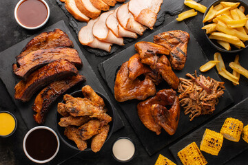 Smoked pork ribs and grilled chicken wings on a slate board, grilled corn and fries on the side on a black background. Various smoked meat set, top view.