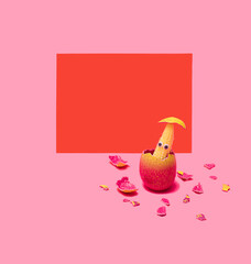 Baby corn with wiggle eyes in pink egg shell with egg shells on the bottom. Red envelope behind. Pink background.