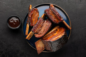 Smoked beef ribs with bone and barbecue sauce on a round dish, top view, rustic black background.