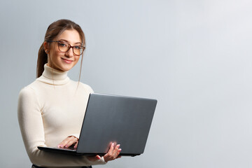 Cute young woman in glasses holds a laptop. Beautiful girl student smiling and looking at the camera on a gray background with copy space.