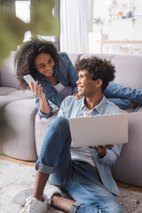 Cheerful african american couple with smartphone and laptop in living room