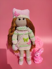 Knitted doll with a rabbit or hare in a dress and a hat with butterflies on pink background. Amigurumi handmade toy