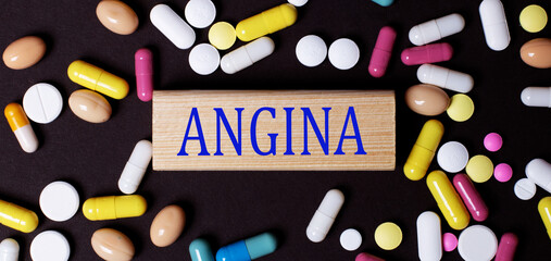 On a dark background, multi-colored pills and the word ANGINA on a wooden block. Medical concept