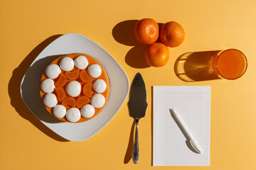 Tangerine cheesecake with meringue discs on white square plate, three tangerines, glass of juice, spatula and notepad for writing a recipe on a yellow table, illuminated by side light