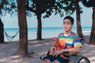 Asian disabled child on wheelchair hug a dinosaur on summer of sea beach nature background,Lifestyle of special child in the education age,Happy disabled kid concept,Toy is best friend in alone mood.