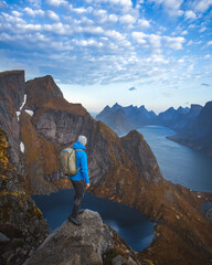 A man with a blue jacket, a hat, and a backpack standing on the top of the mountain, overlooking a beautiful lake and a sea in Lofoten, in Norway during the golden hour.