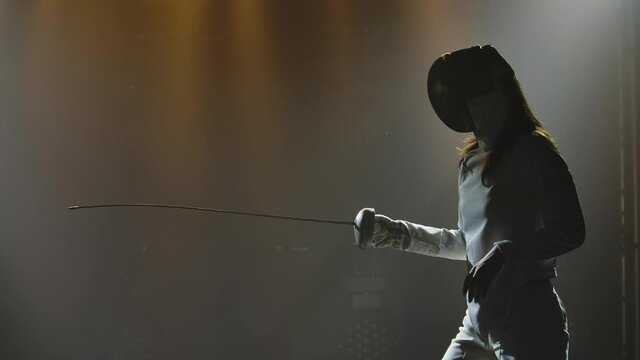 Swordsman woman training with rapier. Female athlete in white uniform and mask practicing fencing exercises and attacking an opponent. Black smoky background in dark studio. Slow motion. Close up.