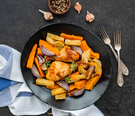 Chicken legs and thighs baked with sweet potatoes or carrots, white and purple onions, regular potatoes on a black ceramic plate on a black concrete background.