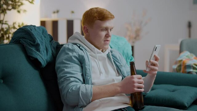 Lustful red-haired man drinking beer, watching adult content video on smartphone