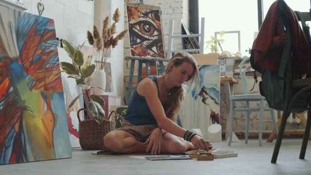 Zoom in shot of young female artist sitting on the floor in studio and drawing picture with crayons