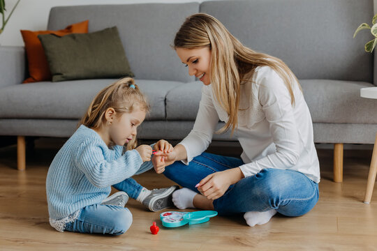 Mother and little daughter paint their nails with toy nail polish. Kid playing with mom at home. Concept of good parenting and happy childhood, family leisure